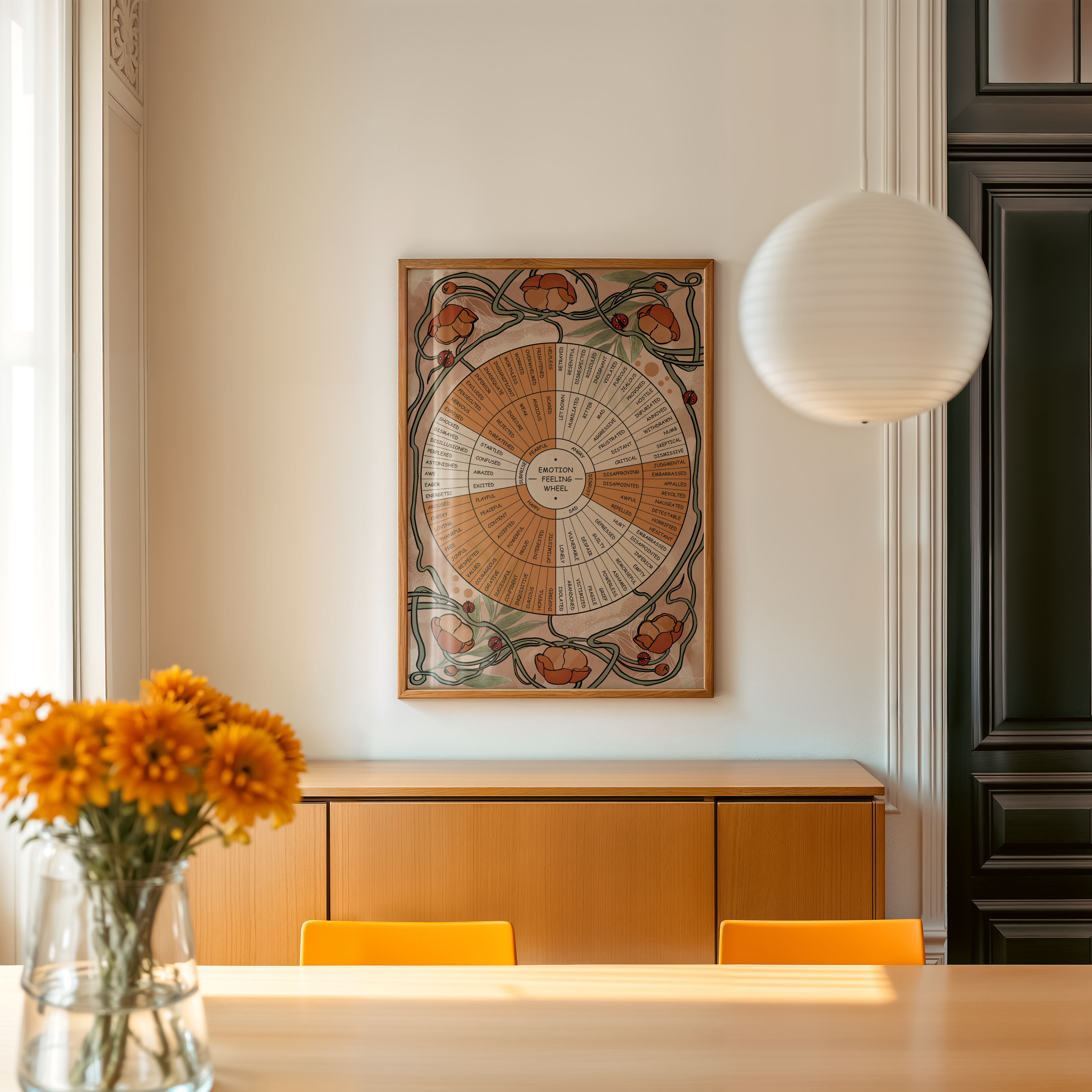 A framed botanical Emotion Wheel poster with illustrated wildflowers, vines, butterflies, and ladybugs hangs above a wooden sideboard in a bright dining room. Yellow dining chairs and a vase of orange flowers create a warm, nature inspired setting.