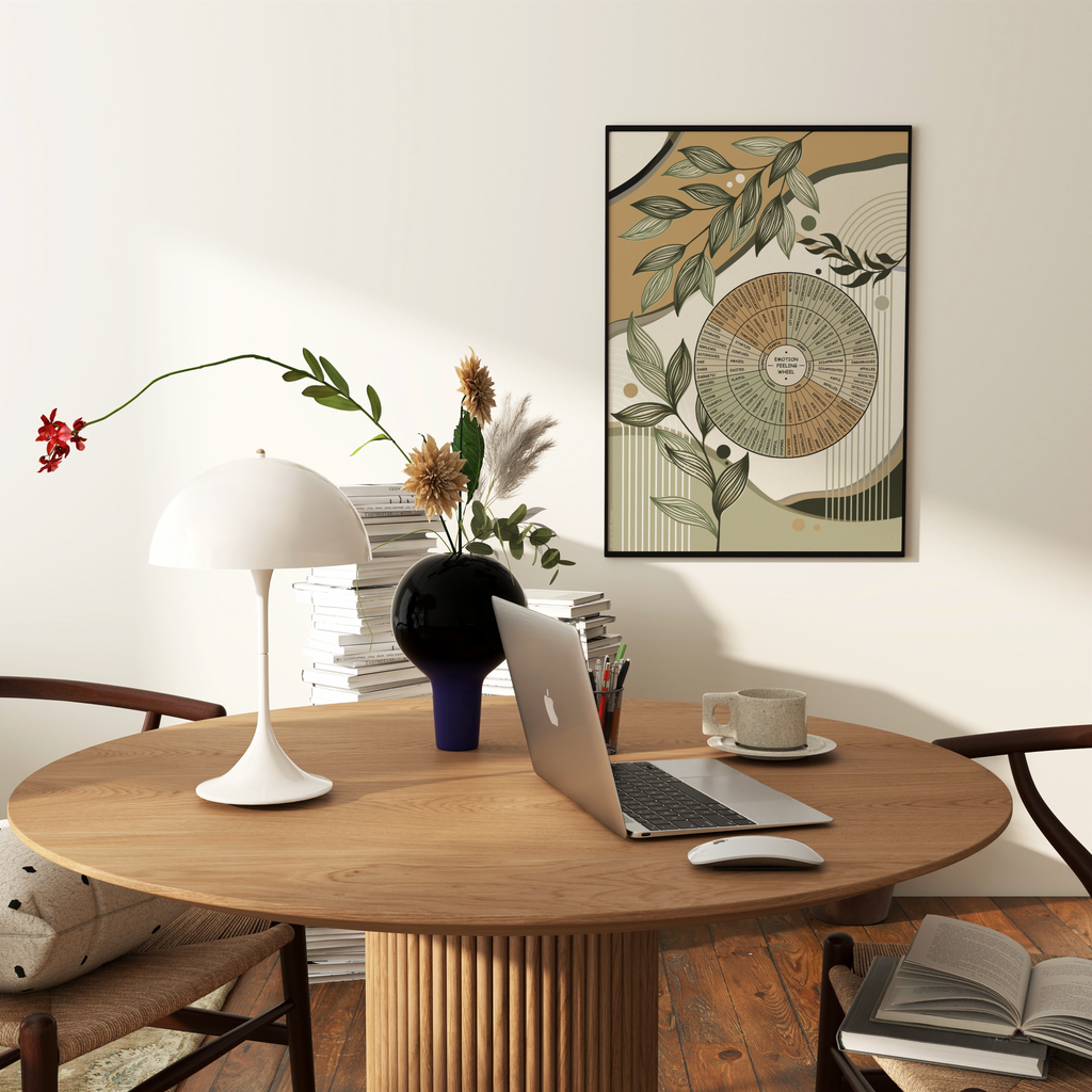 A framed botanical art print hangs on a white wall above a round wooden dining table in a styled home office. The print features a emotion wheel designed with leaves and branches in muted beige, olive green, and brown tones.