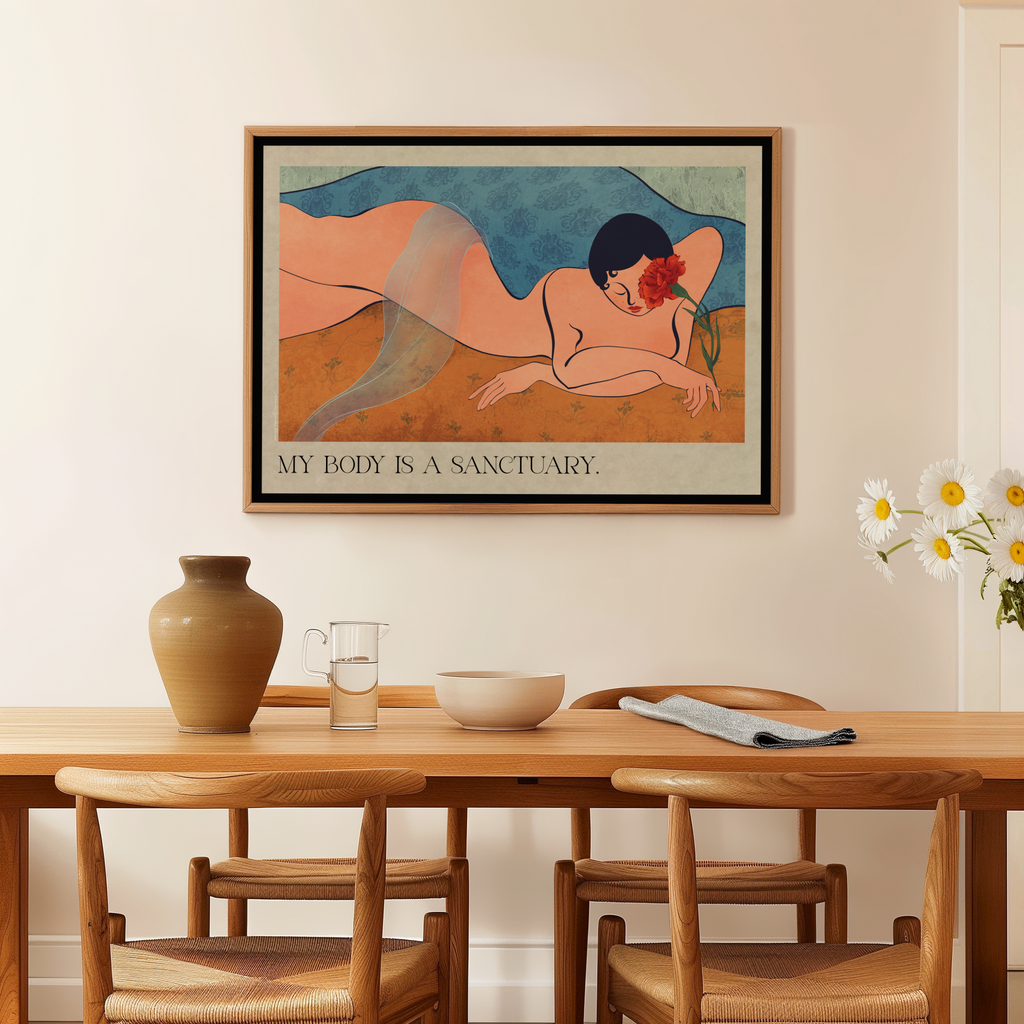 A framed art print featuring a stylized reclining nude figure with the text 'MY BODY IS A SANCTUARY.' displayed on a beige wall above a wooden dining table with chairs, a vase, and a glass.