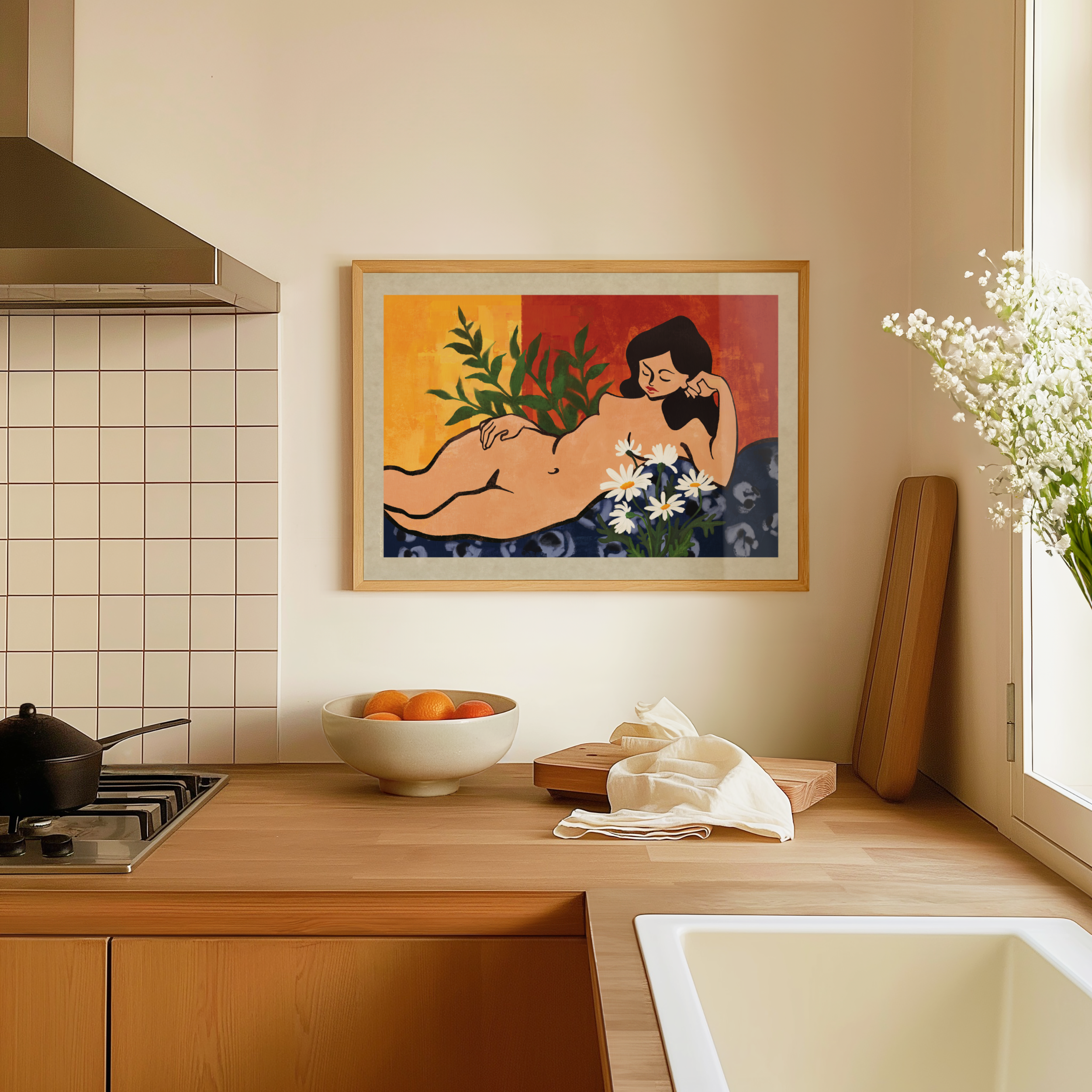 A framed botanical art print featuring a graceful reclining woman surrounded by blooming flowers and lush green leaves hangs on a warm beige wall above a wooden kitchen counter with a bowl of fresh oranges and a nearby sink. The serene artwork celebrates feminine beauty, nature, and self connection, bringing a sense of warmth and tranquility to a cozy, thoughtfully styled home.