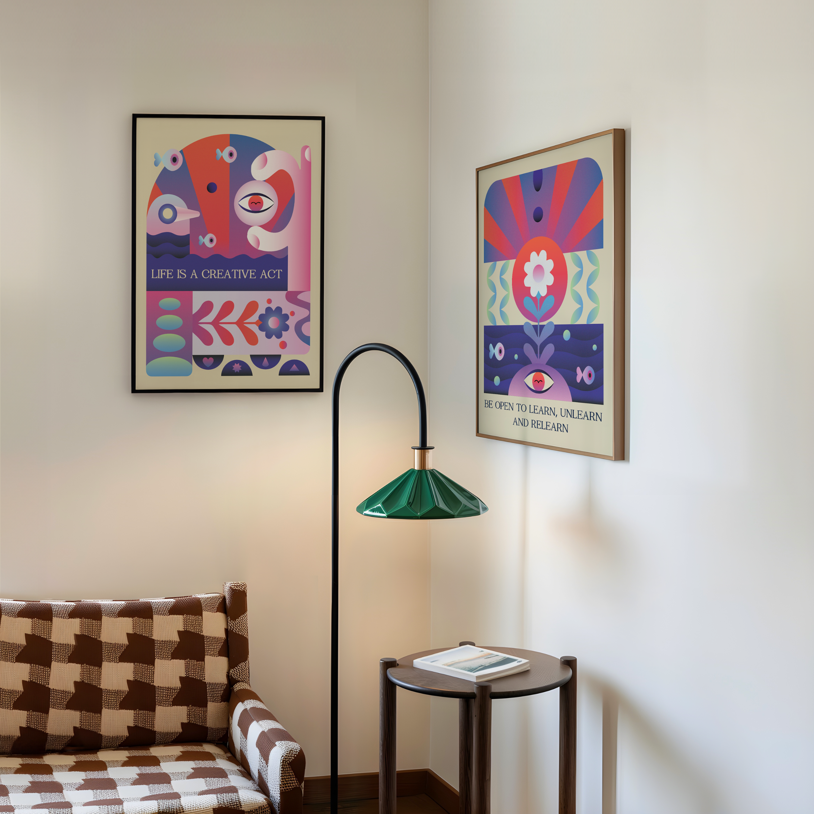 Two framed abstract art prints with colorful geometric and eye-like patterns hang on a white wall in a styled, mid century decorating room. A brown-and-white checkered sofa is visible on the left, a black floor lamp with a green shade stands in the center, and a small round side table with a book is on the right.