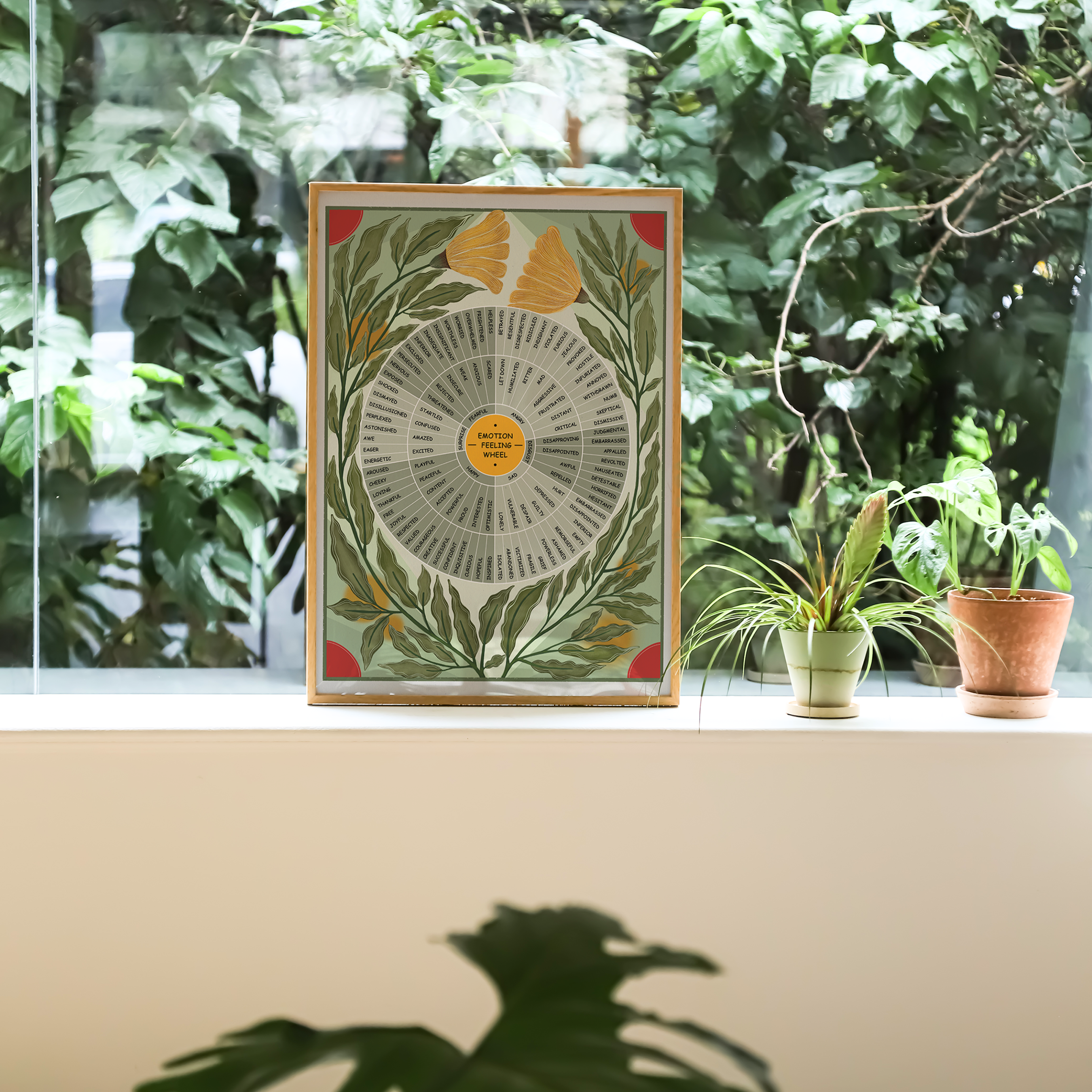 Framed Emotion Feeling Wheel botanical art displayed on a bright windowsill overlooking lush green foliage. The artwork features a detailed emotion wheel surrounded by flowing olive-green leaves and golden ginkgo flowers in a nature-inspired, mid-century modern style. Warm wood framing complements the calming palette, while potted houseplants and soft natural daylight create a peaceful wellness atmosphere. Perfect for therapy offices, counseling spaces, meditation rooms, classrooms, or mindful home decor.