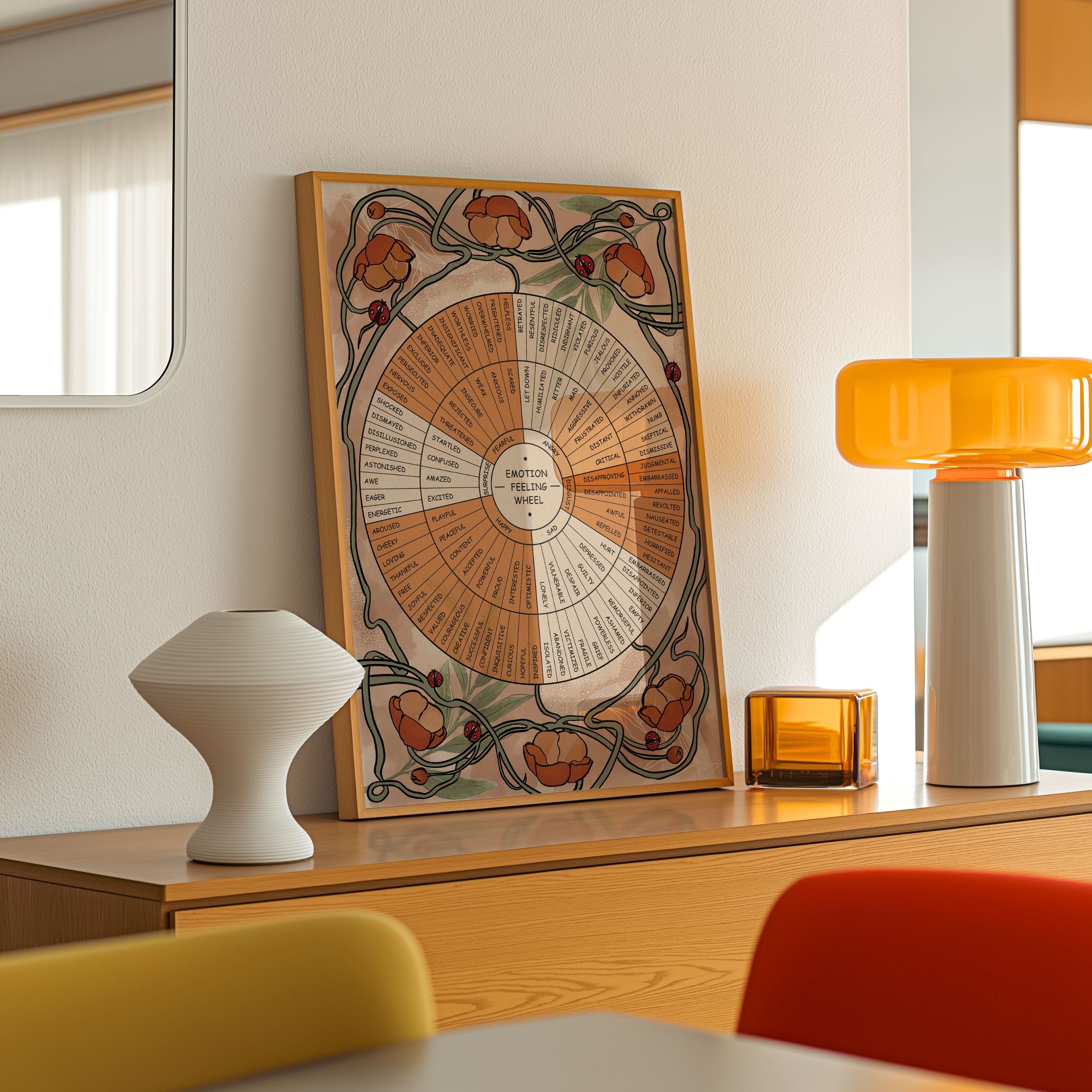 Framed floral Emotion Feeling Wheel artwork displayed on a warm wooden console in a bright mid-century modern interior. The poster features earthy terracotta flowers, winding green vines, and small red ladybugs surrounding a detailed emotion wheel with labeled feelings. Soft natural light highlights the wood frame, ceramic vase, amber glass decor, and orange table lamp, creating a calm, cozy, and mindful space perfect for therapy offices, meditation rooms, or wellness-inspired home decor.