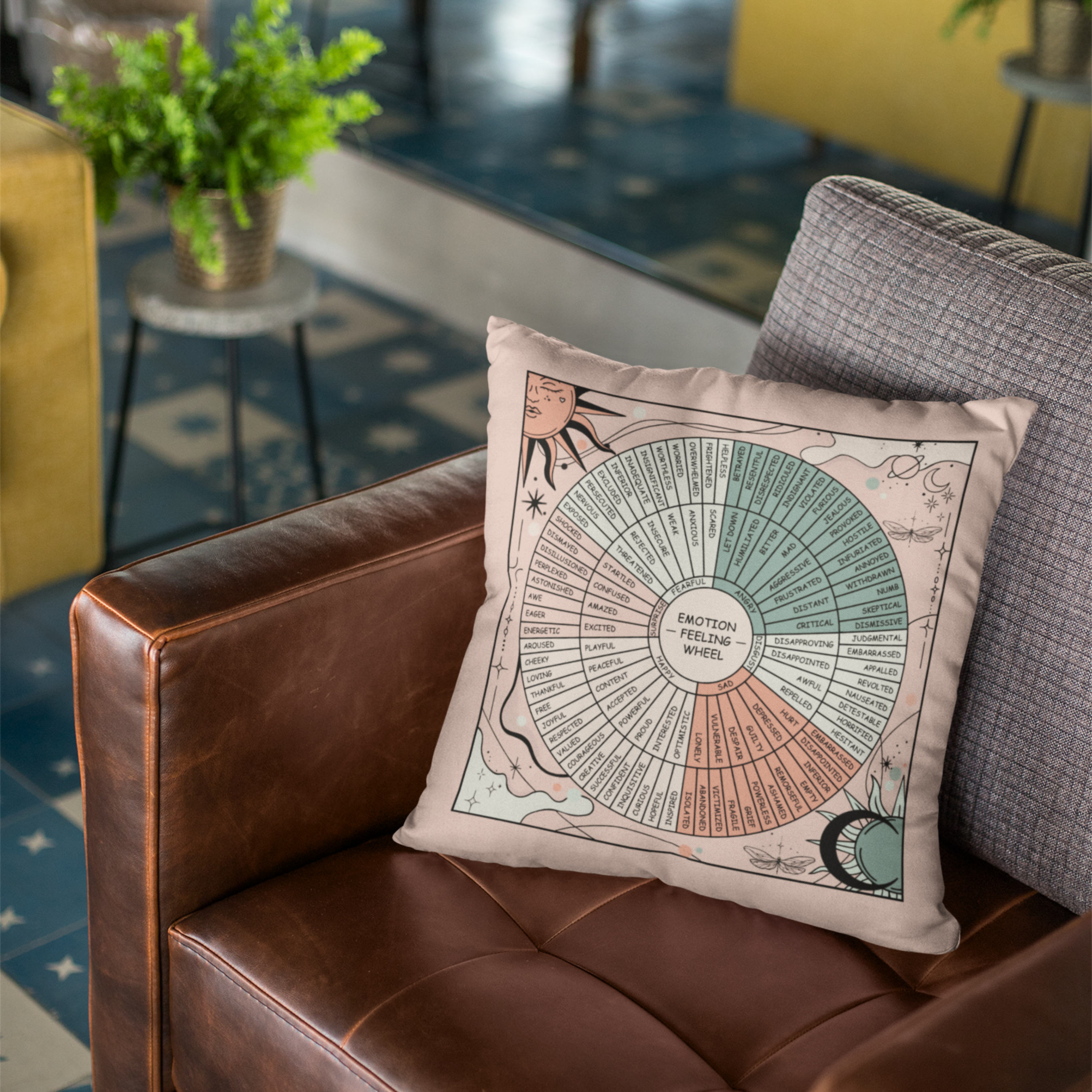 Emotion Feeling Wheel throw pillow displayed on a rich brown leather armchair in a warm, inviting living space. The decorative cushion features a detailed emotion wheel surrounded by celestial illustrations, including the sun, moon, stars, planets, and dragonflies in soft neutral and pastel tones. Cozy textures, modern furnishings, and greenery create a calming atmosphere, making this mental health pillow ideal for therapy offices, counseling rooms, reading nooks, meditation spaces, or mindful home decor.