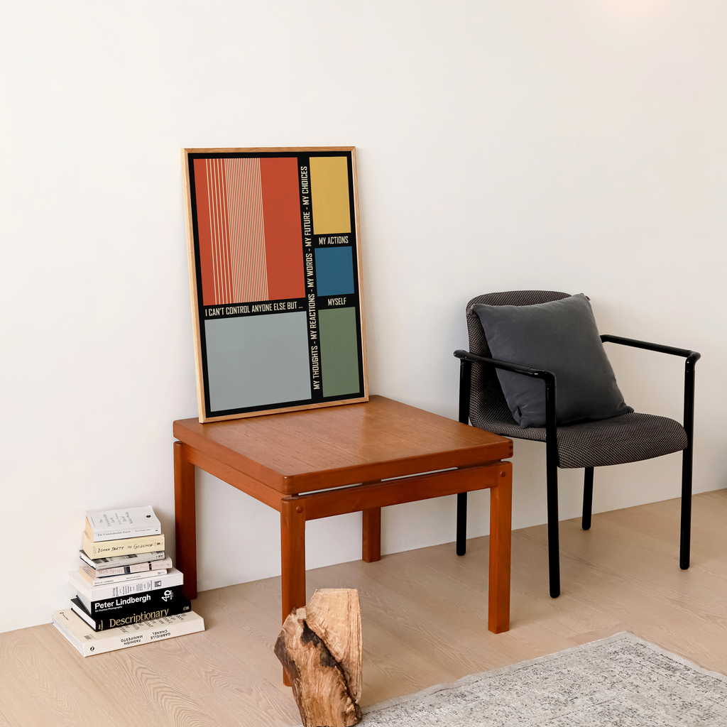 A framed abstract geometric art print featuring vertical color blocks in red, orange, yellow, blue, green, and gray leans against a white wall on a wooden side table. Styled beside a gray upholstered armchair with a dark gray cushion and a stack of books, the modern artwork brings bold color, clean geometry, and a creative focal point to a cozy living room, reading nook, or contemporary interior.