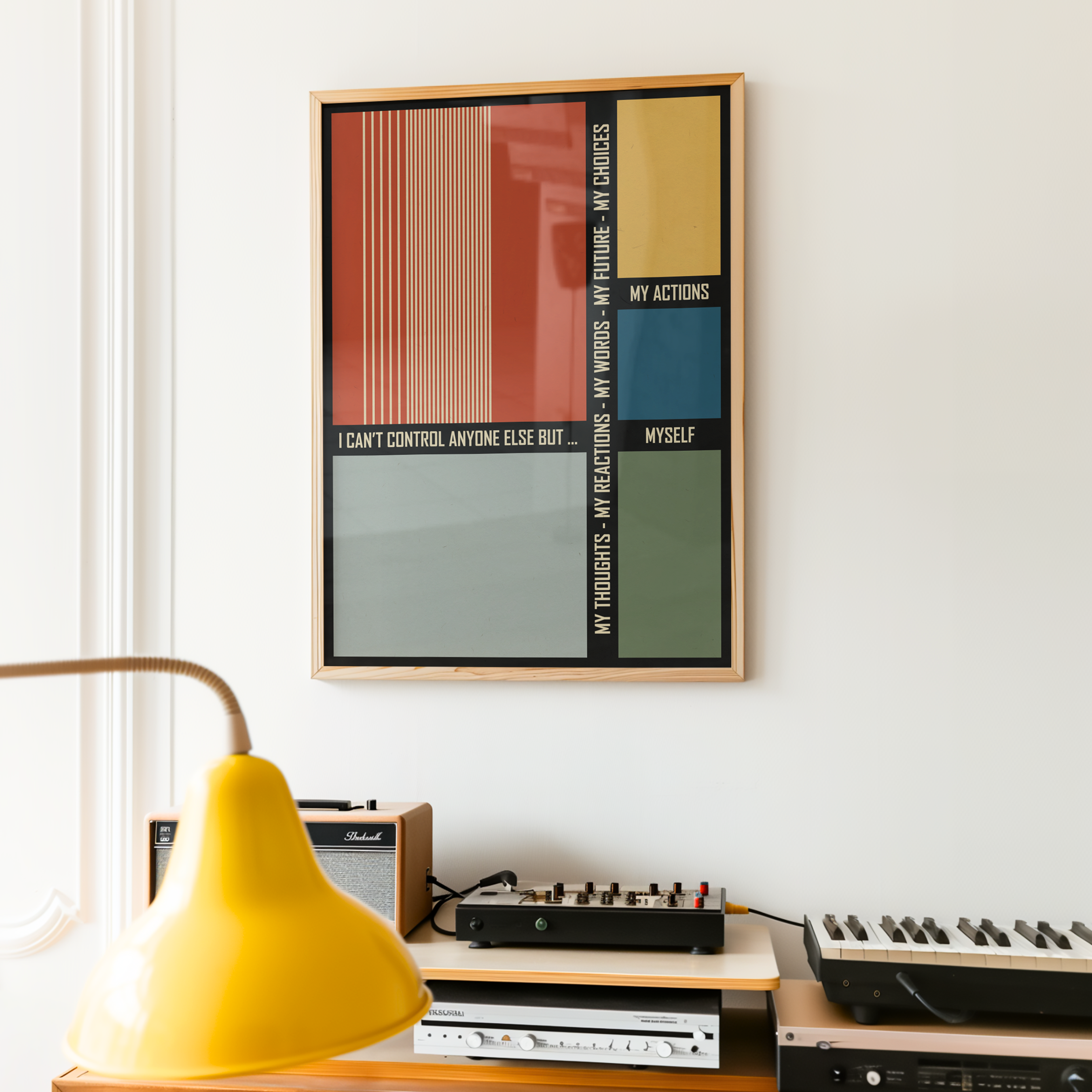 A framed motivational typography poster featuring a bold grid of colorful geometric blocks and inspirational text hangs on a white wall above a modern desk with a yellow table lamp, keyboard, and amplifier. The contemporary, mid-century, bauhaus-inspired artwork brings creativity, focus, and positive energy to a home office, studio, or workspace while adding a striking modern design element.