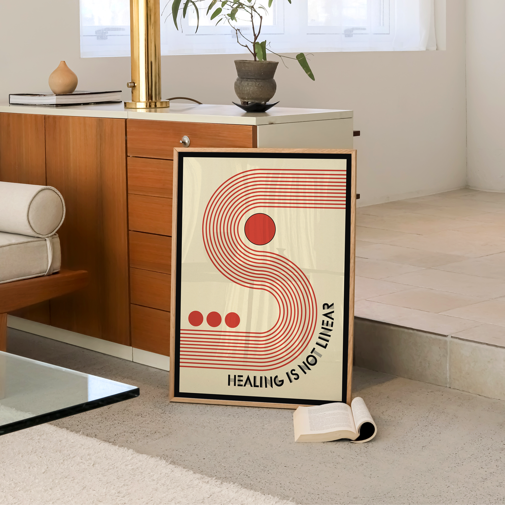 A framed Healing Is Not Linear typography art print with a bold retro inspired design leans against a wooden cabinet in a modern living room. Featuring a striking red letter S, geometric shapes, and uplifting typography, this motivational wall art celebrates the healing journey with warmth and encouragement, making it a meaningful addition to therapy offices, counseling spaces, wellness studios, or mindful home decor.