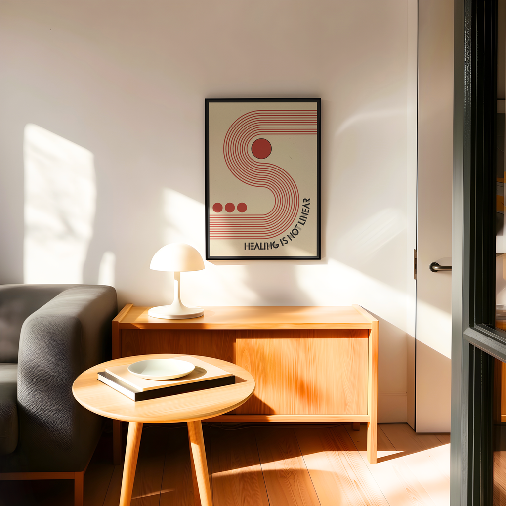 A framed Healing Is Not Linear art print featuring a retro red and beige wavy design hangs on a white wall above a wooden sideboard in a bright, sunlit living room. Inspired by mid century modern and Bauhaus design, the artwork combines bold typography with geometric forms to create a meaningful reminder that healing is a journey. Styled with a gray armchair, a round wooden table, and a white lamp, it brings warmth and inspiration to therapy offices, home offices, or contemporary living spaces.
