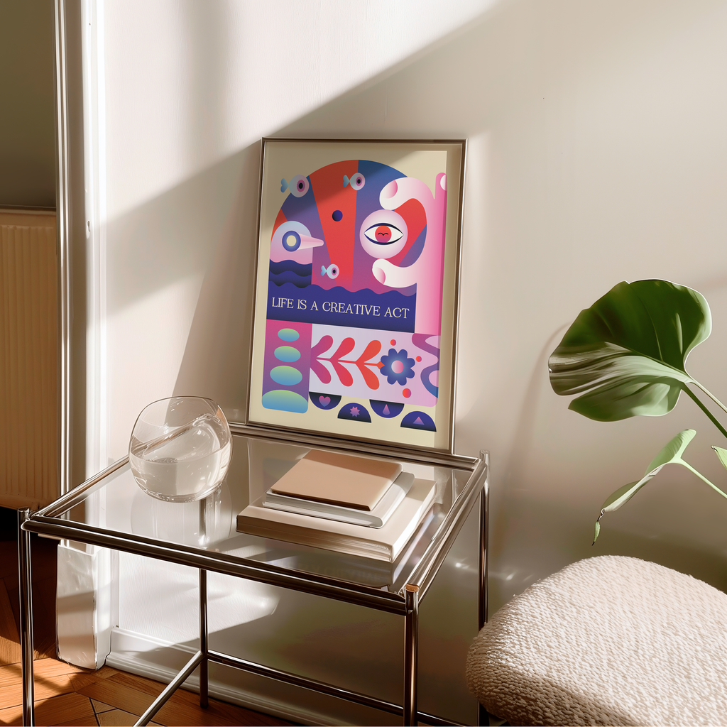 A framed Life Is a Creative Act art print leans on a glass side table in a bright, sunlit room beside a stack of books, a clear glass bowl, and a lush green plant. Inspired by mid century modern and Bauhaus design, the artwork features bold geometric shapes and vibrant colors that celebrate creativity, imagination, and self expression, adding an uplifting focal point to a modern living space, studio, or home office.