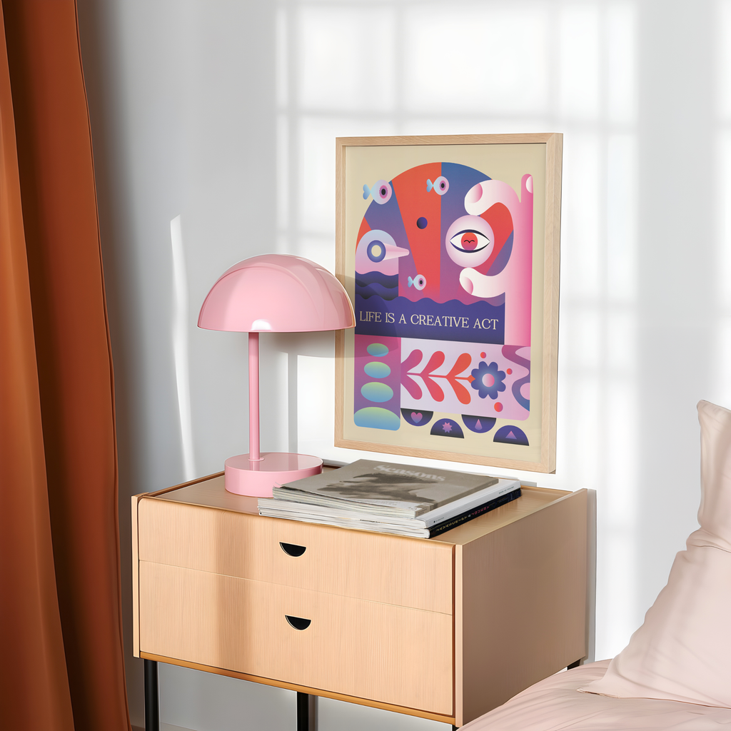 A framed Life Is a Creative Act art print is styled on a light wood nightstand beside a pink table lamp and a stack of books in a cozy bedroom. Inspired by mid century modern and Bauhaus design, the artwork features colorful abstract faces, geometric shapes, and expressive typography that celebrate creativity, self expression, and everyday inspiration, bringing a playful yet thoughtful accent to modern interiors.