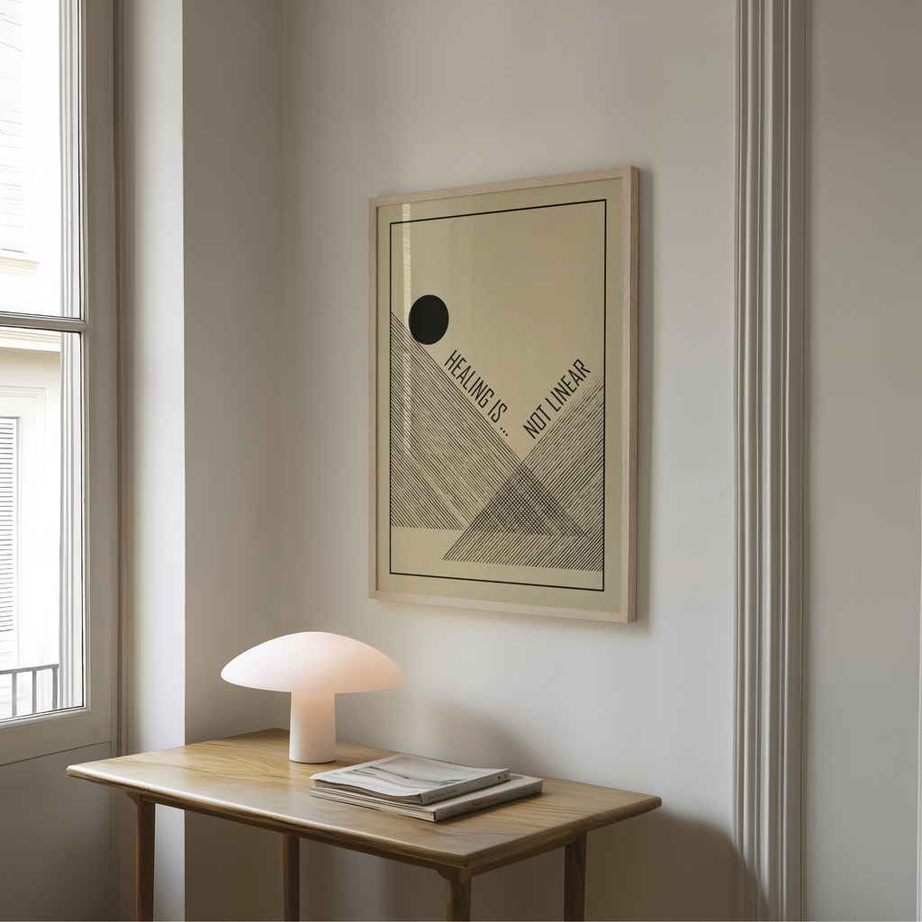 A framed Healing Is Not Linear art print featuring minimalist black geometric line art and typography on a cream background hangs on a light gray wall above a wooden table with a mushroom shaped lamp and stacked books. Inspired by mid century modern and Bauhaus design, the artwork combines clean forms and a meaningful message, offering a gentle reminder that healing is a personal journey with its own rhythm. Perfect for therapy spaces, wellness rooms, home offices, or mindful interiors.