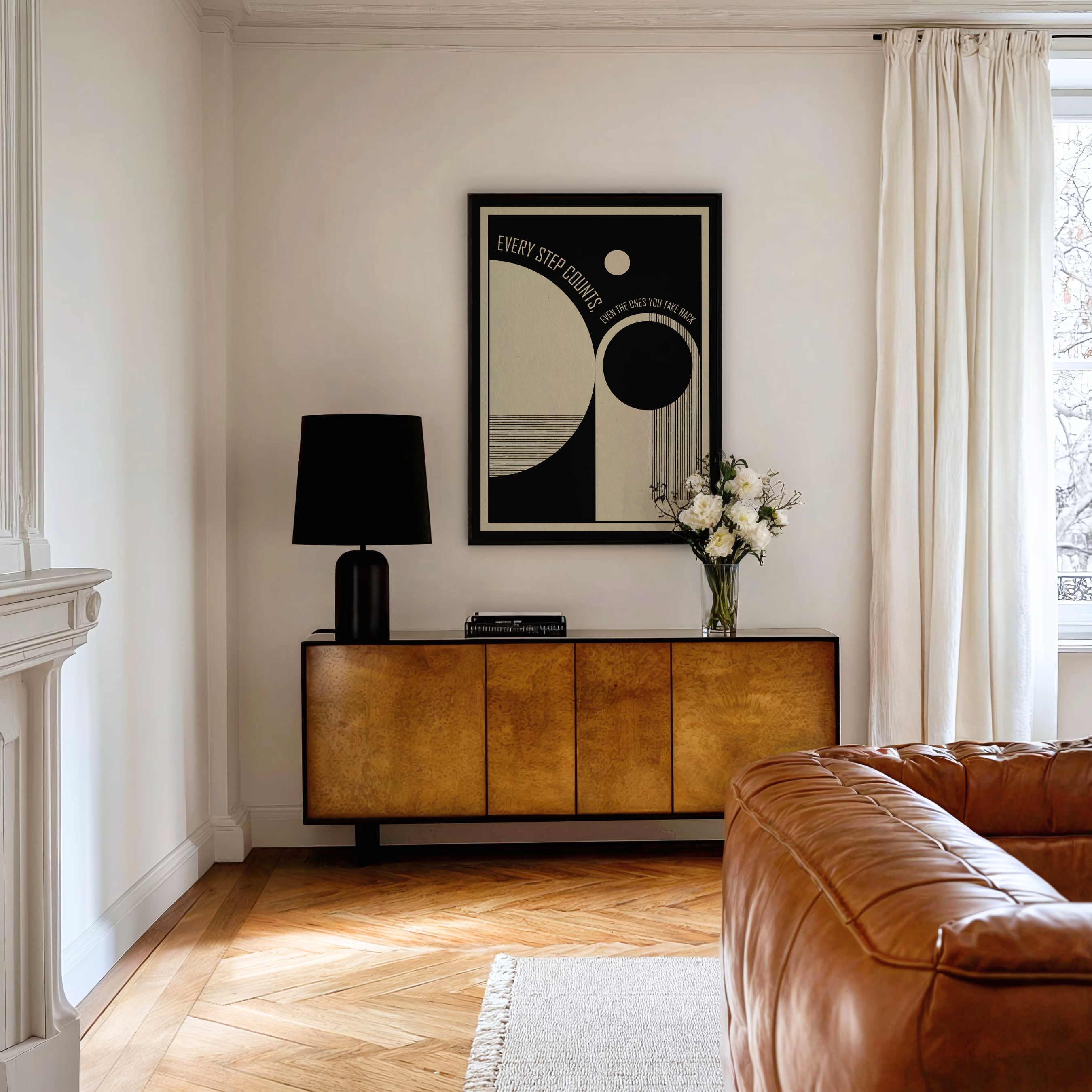 A framed Every Step Counts, Even the Ones You Take Back art print featuring black, white, and beige geometric shapes hangs on a white wall above a warm wood cabinet in a thoughtfully styled room. Inspired by mid century modern and Bauhaus design, the artwork combines clean lines, balanced forms, and a meaningful message about growth, resilience, and the healing journey. Styled with a brown leather sofa, black table lamp, and vase of white flowers, it brings a calm and reflective touch to modern interiors.