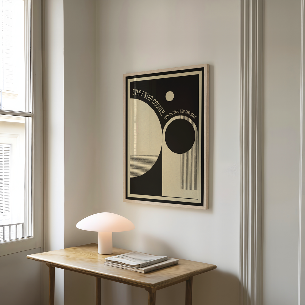 A framed Every Step Counts, Even the Ones You Take Back art print featuring minimalist black and cream geometric shapes hangs on a light gray wall above a wooden table with a glowing mushroom shaped lamp and stacked books. Inspired by mid century modern and Bauhaus design, the artwork blends clean lines, balanced forms, and an encouraging message about growth and resilience, creating a calm and thoughtful accent for modern homes, therapy spaces, home offices, or mindful living areas.