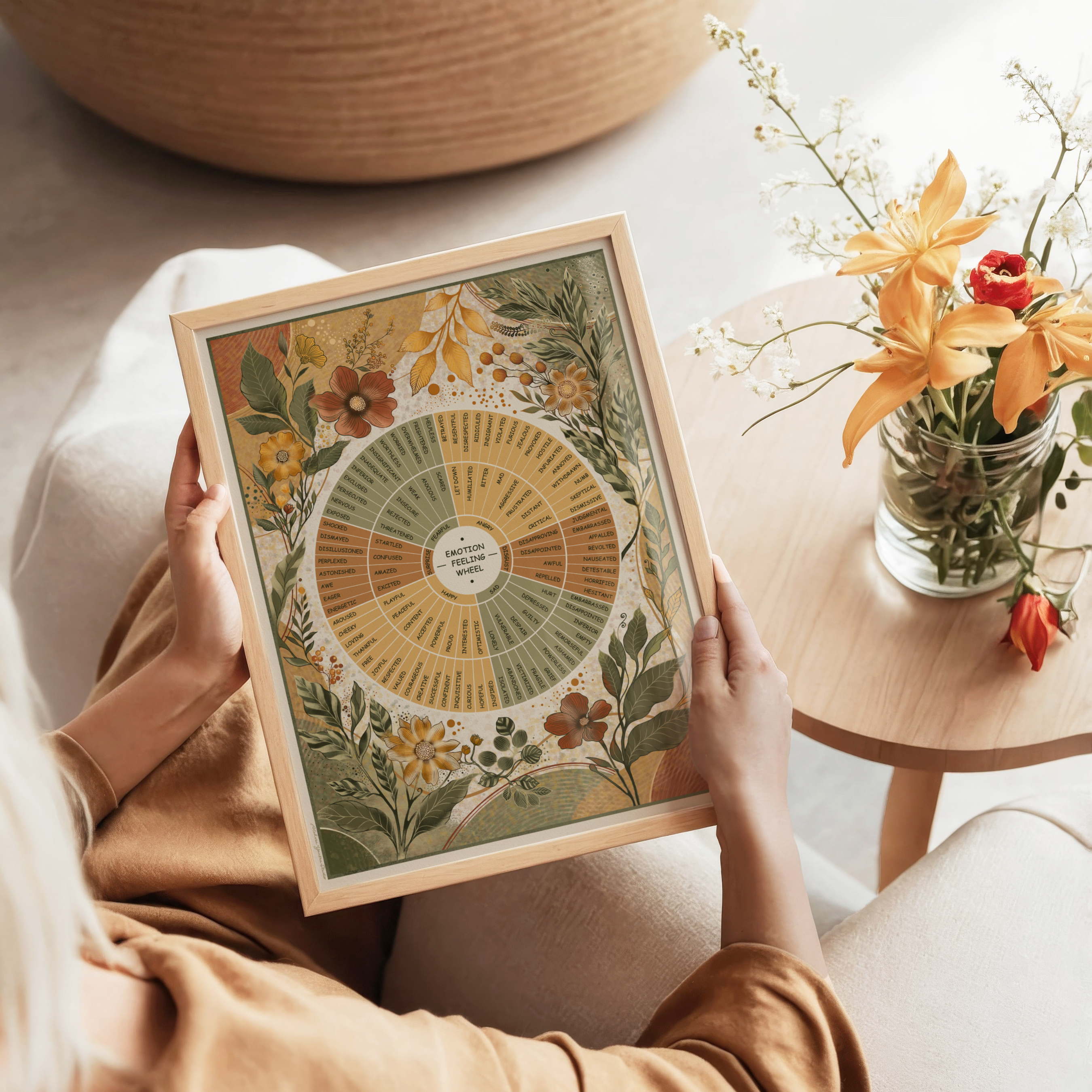 A person seated indoors holds a framed botanical infographic print. The artwork features a therapy chart surrounded by illustrated flowers and leaves in muted yellow, green, orange, and brown tones.