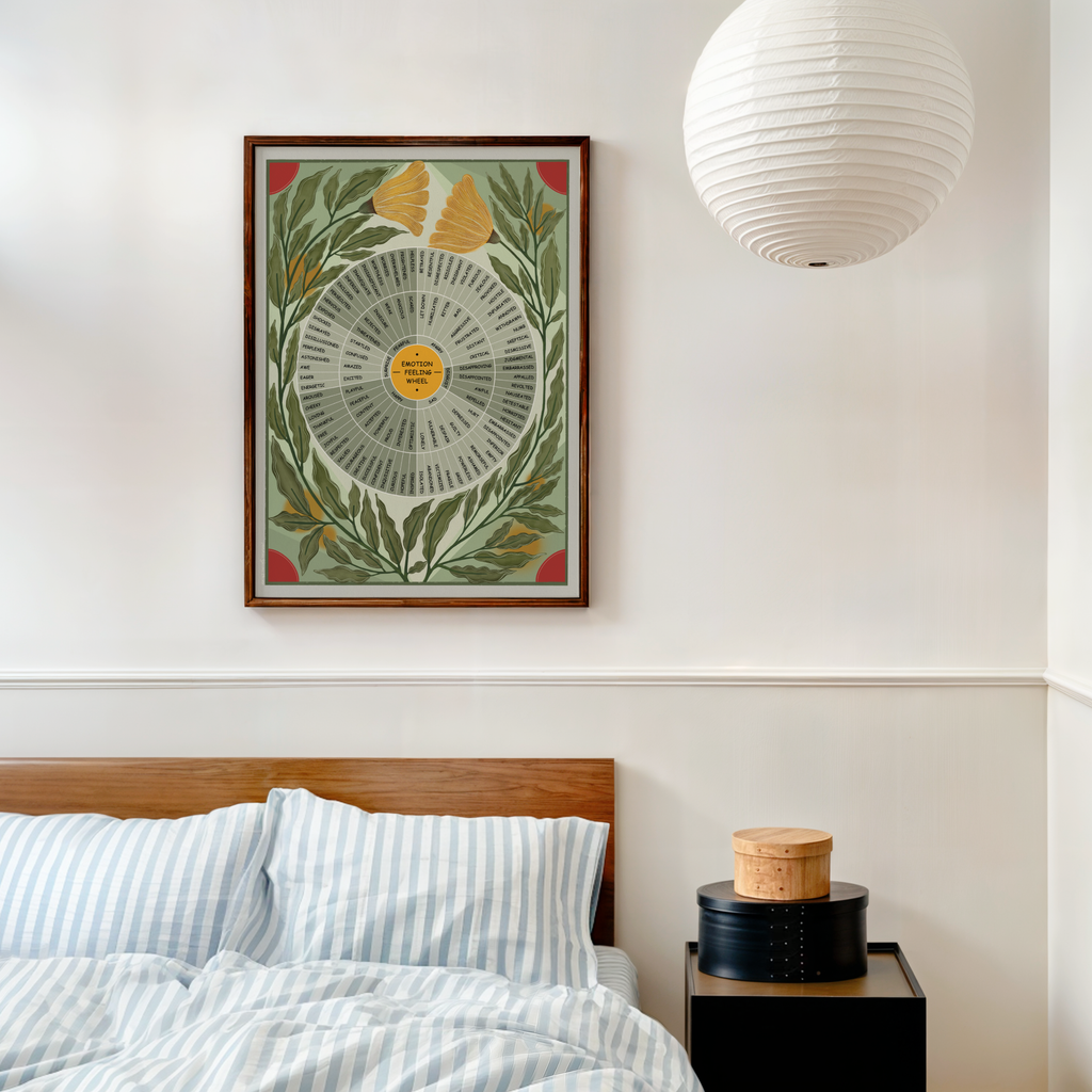 A framed vintage-style botanical art print with a feelings wheel hangs on a white wall above a bed with blue-and-white striped bedding. A white paper lantern hangs to the right, and a small bedside table holds stacked round boxes.