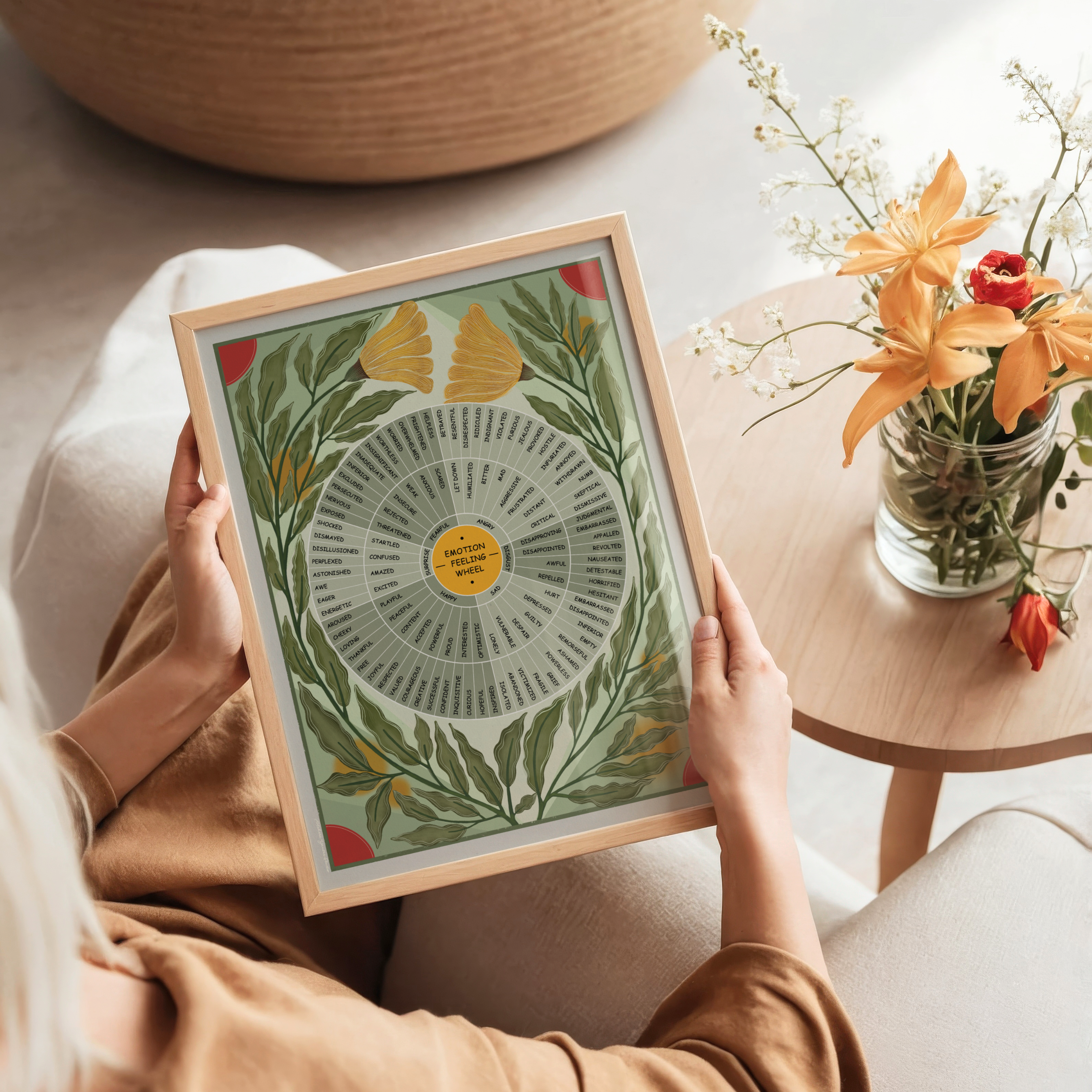 A person holds a framed botanical-style emotion wheel with green leaves, yellow accents, and a gold center label. The scene is styled in a cozy indoor setting with a wooden side table holding orange flowers in the background.