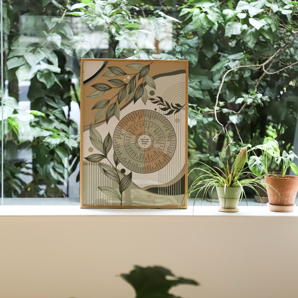 A framed botanical art print with an emotion wheel illustrations, displayed on a windowsill with potted plants and a lush green outdoor background.