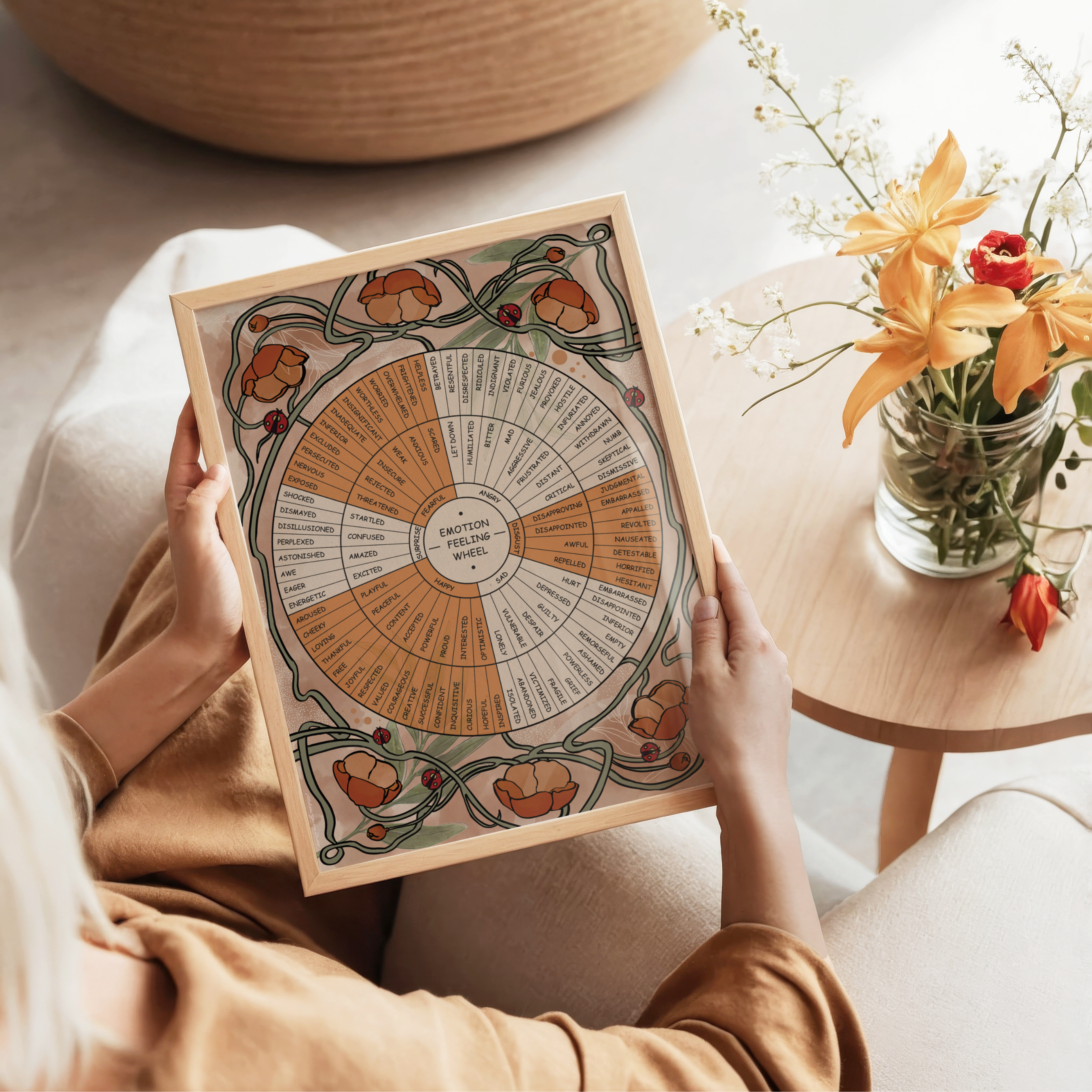 A person holds a framed Wildflower Emotion Wheel poster featuring hand drawn botanical illustrations, delicate wildflowers, butterflies, and ladybugs in a cozy indoor setting. A small wooden table with a vase of orange flowers complements the warm, nature inspired artwork.