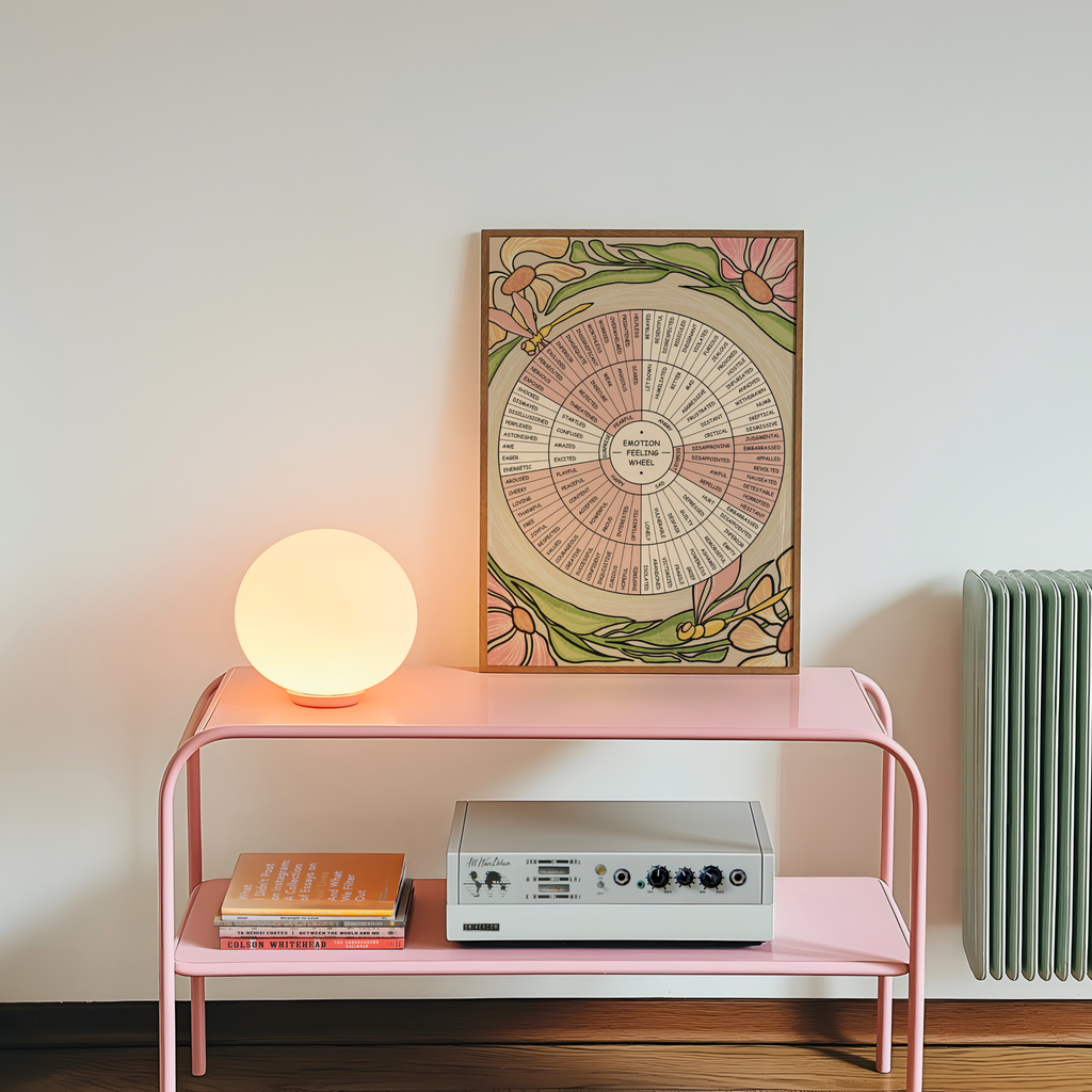 A framed vintage-style floral emotion wheel print hangs on a light wall above a pink metal shelf. A glowing round lamp sits on the top shelf, with books and a silver audio device on the lower shelf.