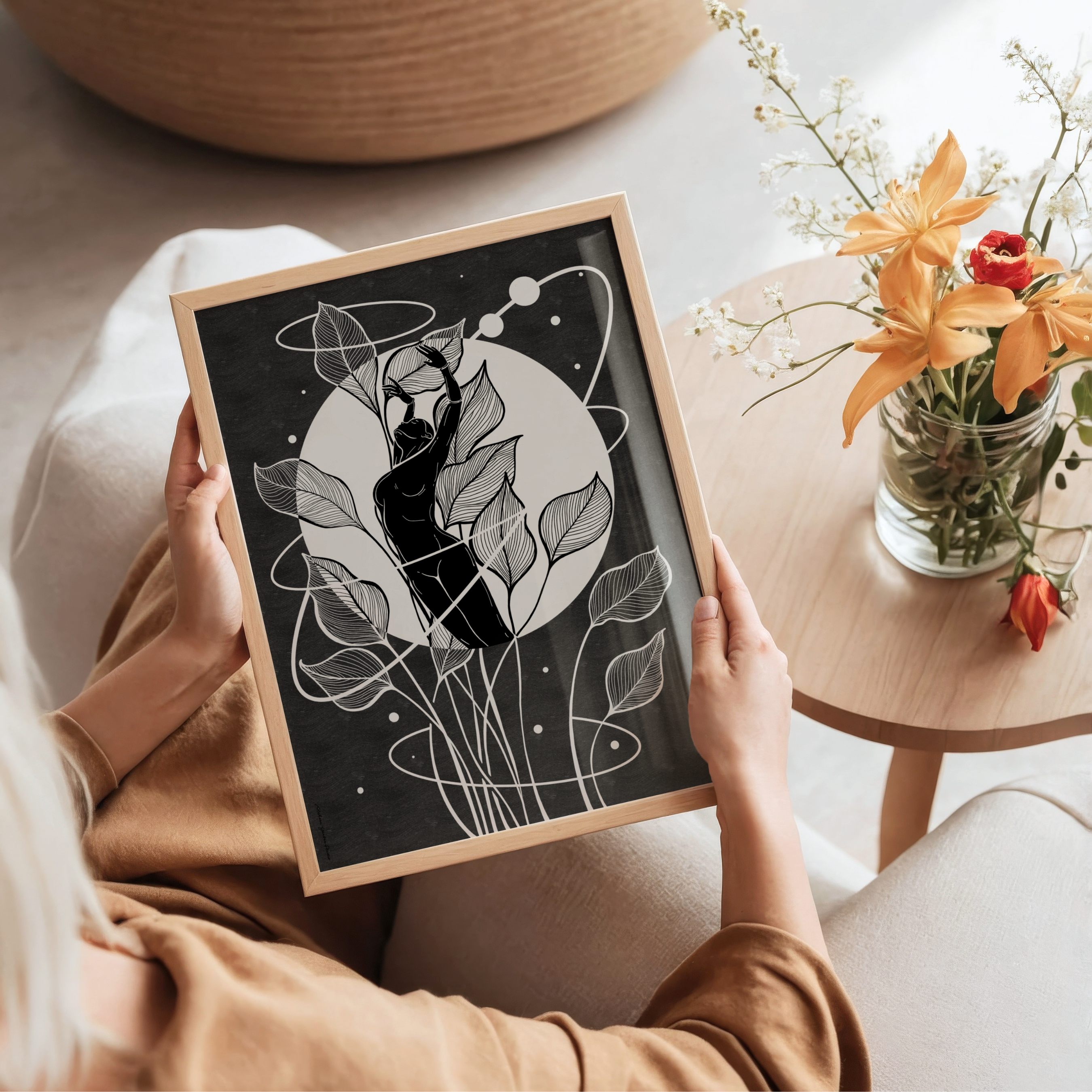 A person holds a framed black and white art print featuring a minimalist illustration of a feminine figure beneath a full moon with flowing organic line art. Inspired by the menstrual cycle and the lunar cycle, this self love artwork celebrates feminine wisdom, inner balance, and the natural rhythms of the body in a cozy indoor setting with a wooden side table and fresh flowers nearby.