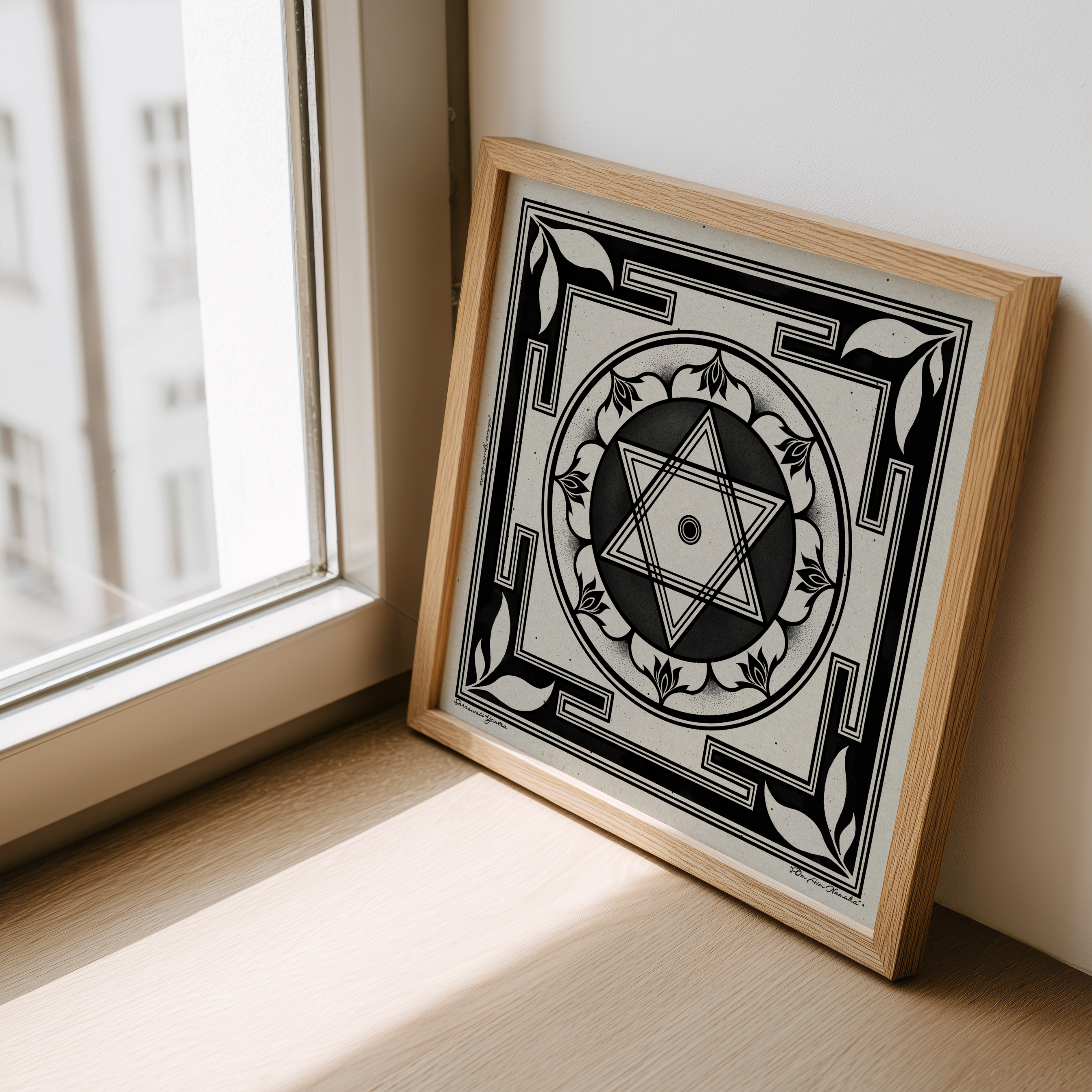 A square framed art print featuring a black and white geometric Saraswati yantra, leaning against a wall on a windowsill in a sunlit room.