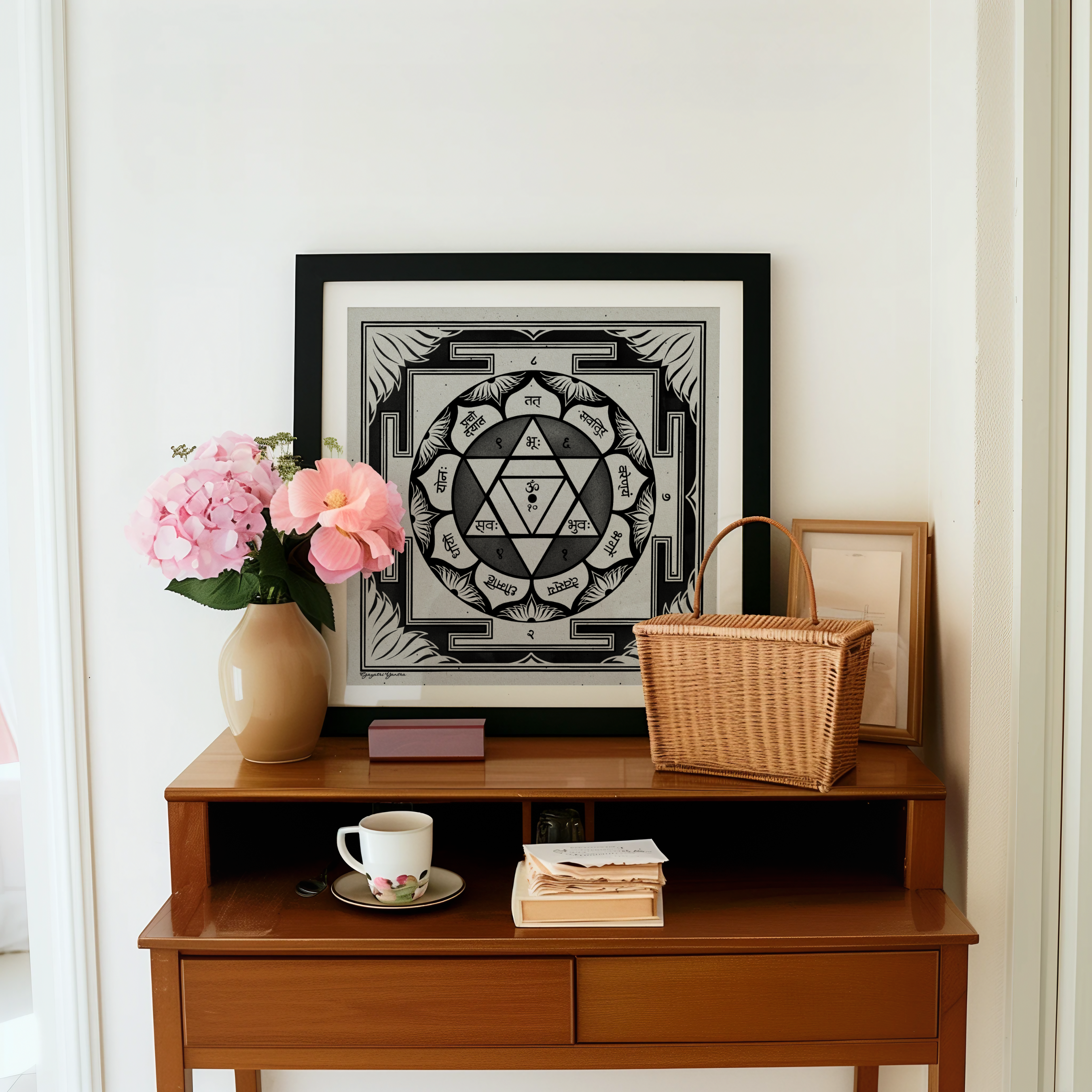 A framed black and white Gayatri yantra art print with a yellow triangle in the center, displayed on a wooden console table in a styled room setting with pink flowers, a wicker basket, and a white mug.