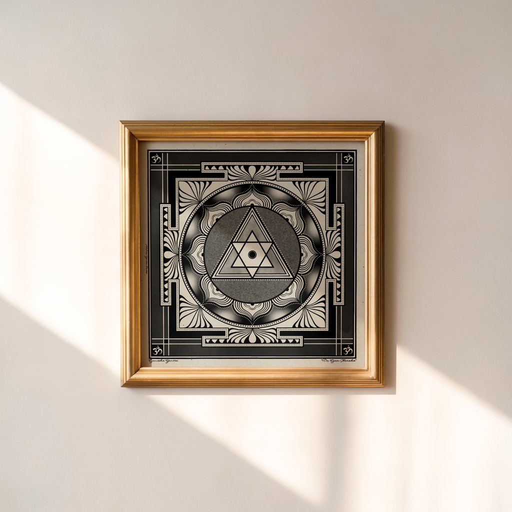 A framed square art print featuring a black and white Ganesha yantra with Sanskrit script, housed in a gold frame and mounted on a white wall with soft diagonal sunlight shadows.