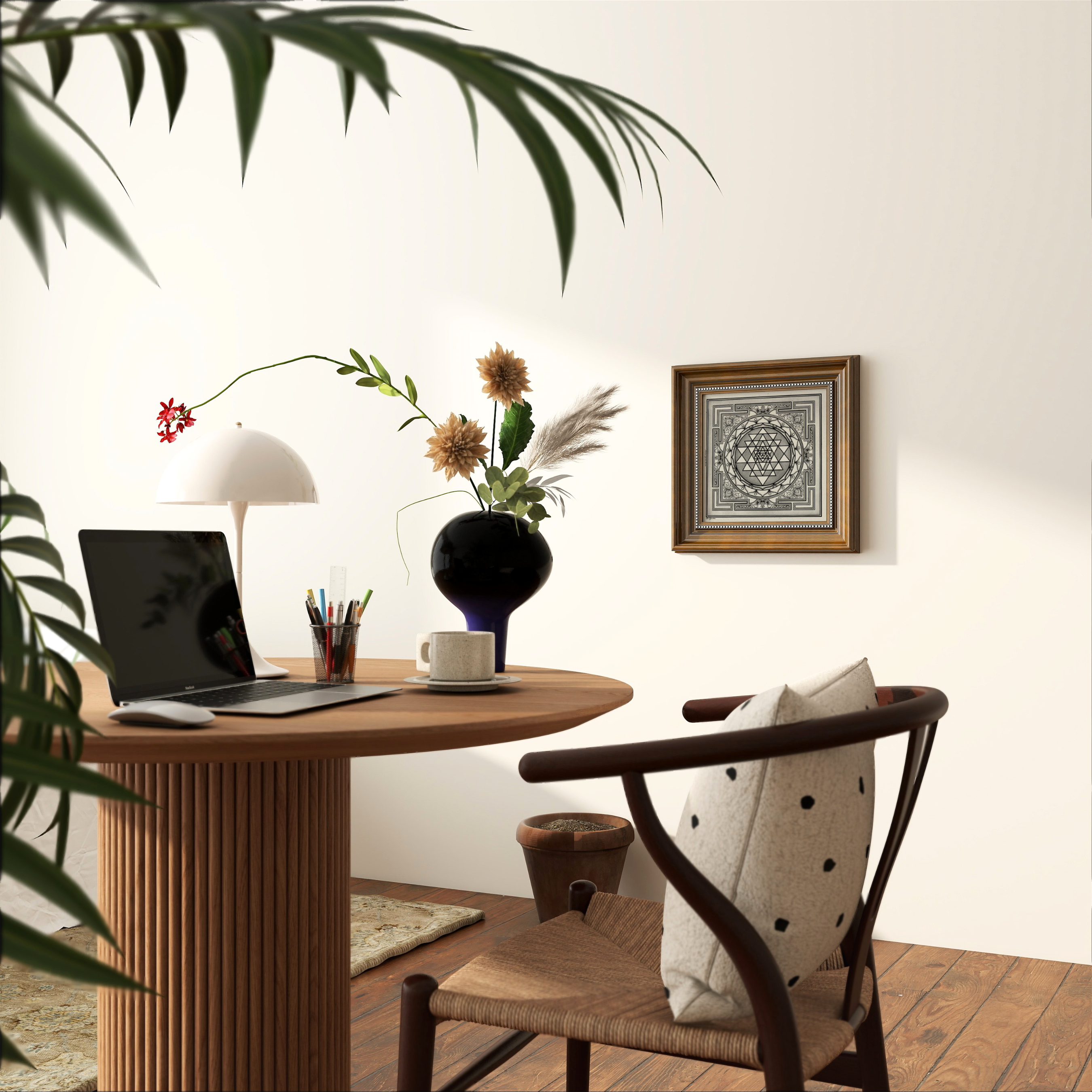 A styled home office corner featuring a round wooden desk with a ribbed pedestal base, a laptop, a white dome lamp, a black vase with dried flowers, and a woven wood chair with a cream pillow. A framed mandala Sri yantra print hangs on the off-white wall, with a leafy plant in the foreground and warm sunlight casting shadows.