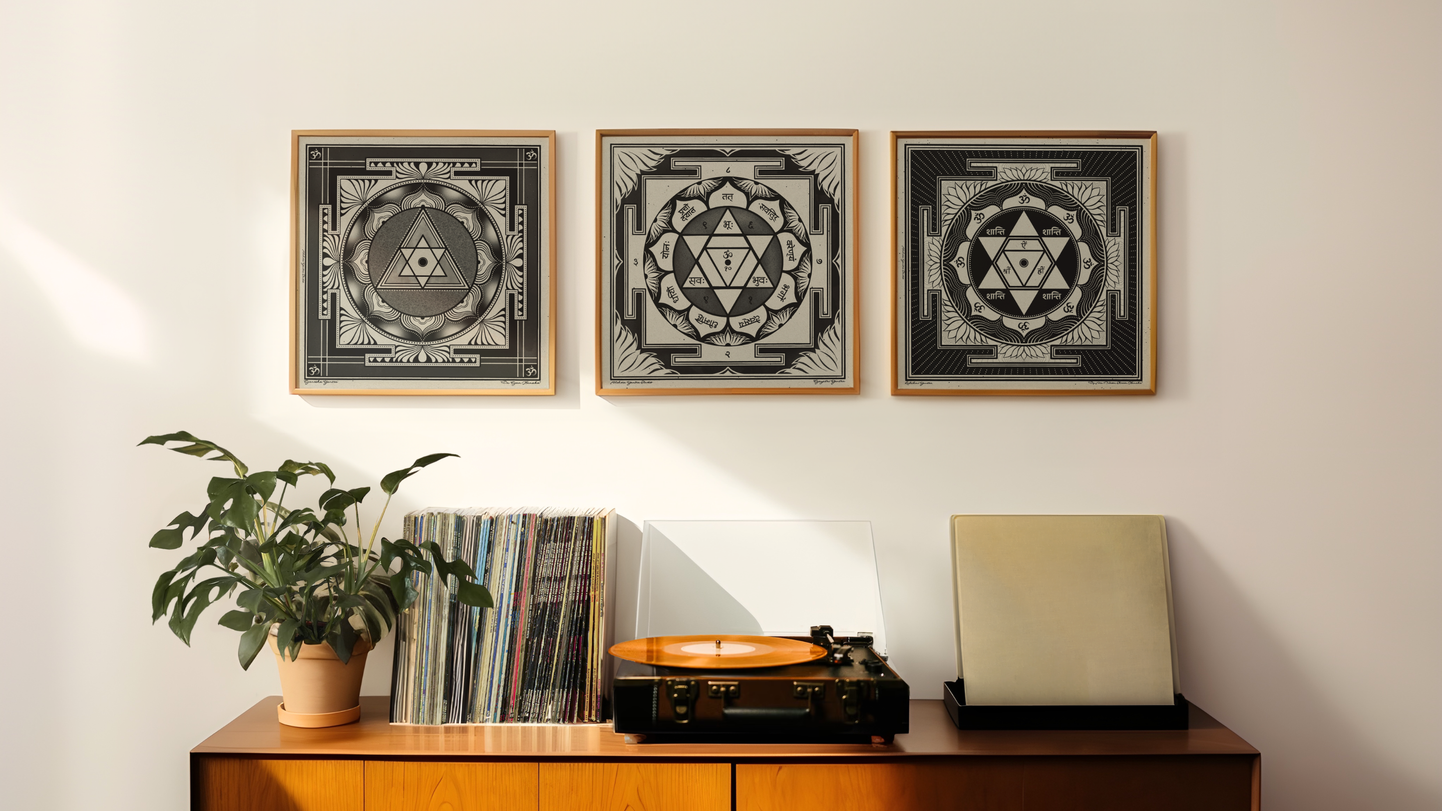 Three framed black and white mandala art prints with Sanskrit mantras are displayed on a white wall above a wooden console table with a plant, vinyl records, a turntable, and a speaker.